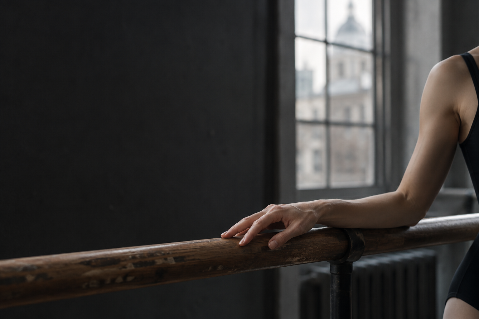 A dancer’s hand resting on a wooden ballet barre in cool studio light